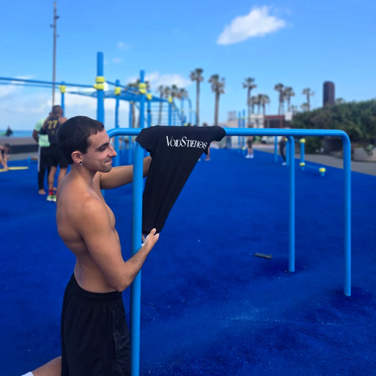 Un atleta de VoidSthenos sujeta una camiseta de la escuela sobre la barra de un parque de calistenia junto al mar.